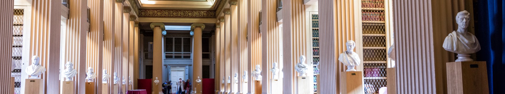 A grand, classical interior hall with tall fluted columns, an ornate arched ceiling, and a long red carpet, lined with marble busts on pedestals leading toward a distant doorway