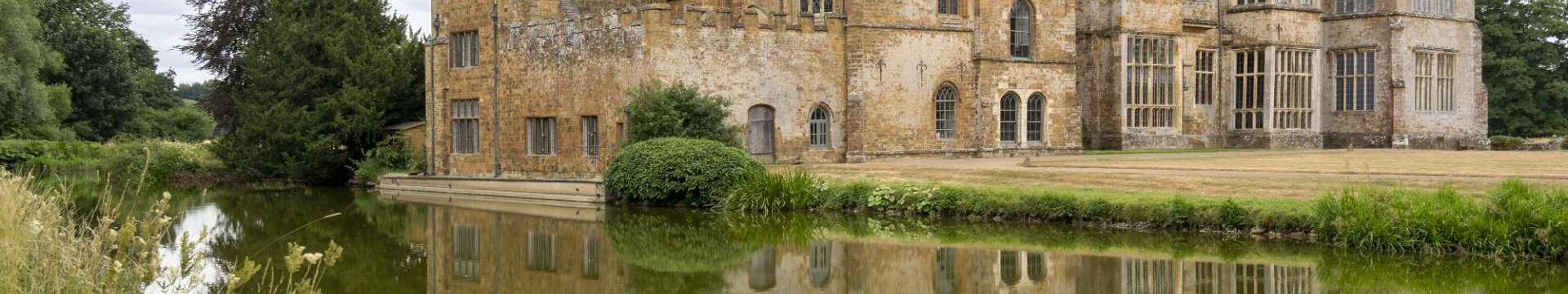Historic stone manor house beside a calm moat, reflected in the water under a cloudy sky, with trees and grasses surrounding the grounds
