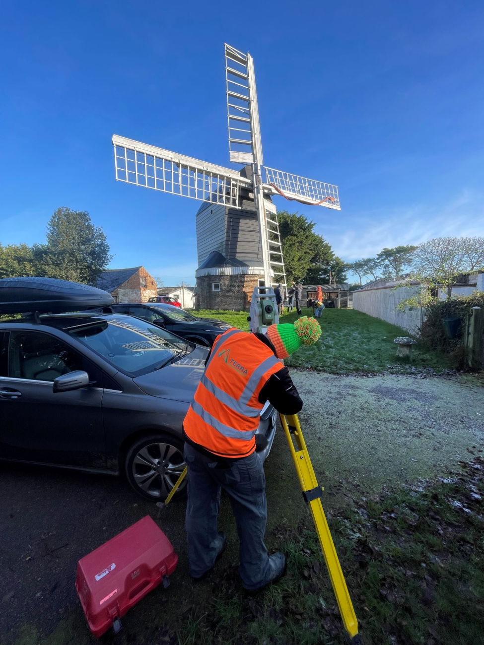 A man in a hi-viz vest uses a surveyors machine in front of a windmill