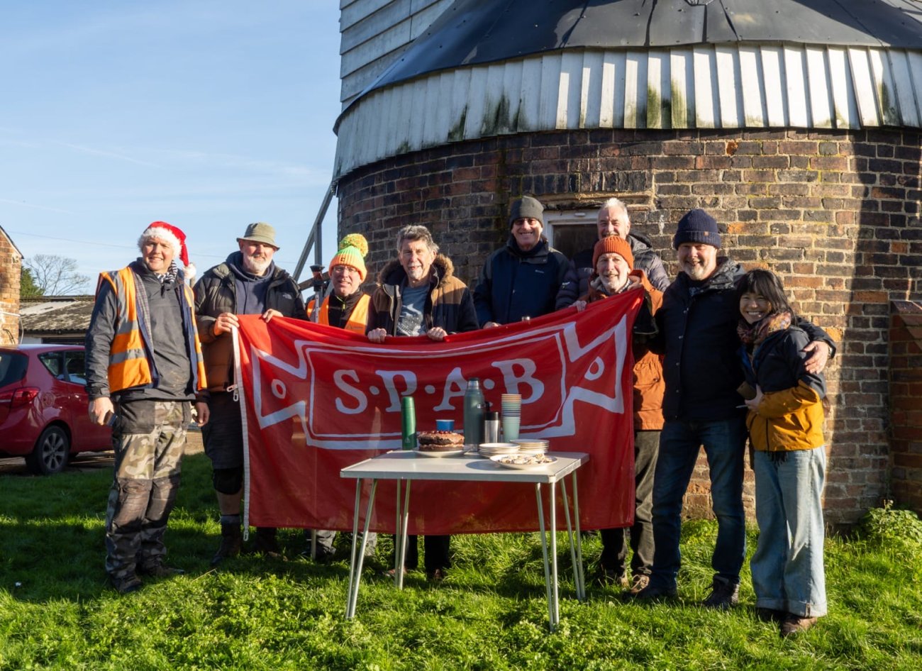 A group of people gathered in front of a windmill base with a SPAB Banner