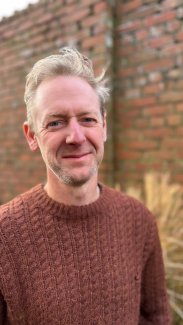 Matt James looking at the camera. Man with grey hair and a brick wall behind