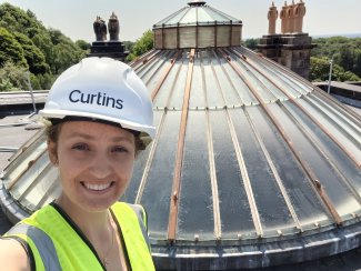 A woman stands in front of a glass roof dome. She's smiling at the camera and wearing a hard hat.
