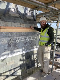 A smiling man with a beard stands on scaffolding in front of historic details. He has a hard hat and high-vis vest.
