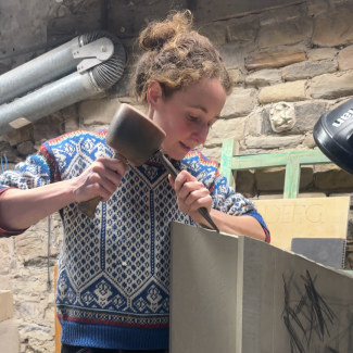 Grace Impesi working at stone masonry in a workshop. Using a hammer and chisel on a stone.