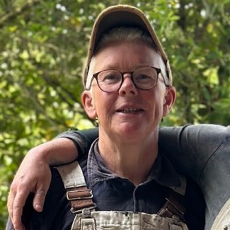 Han Bee looking at the camera. They are wearing glasses, a cap and dungarees with a tape measure attached.