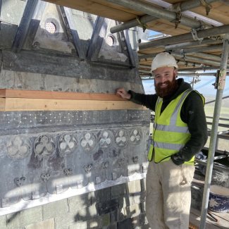 A smiling man with a beard stands on scaffolding in front of historic details. He has a hard hat and high-vis vest.