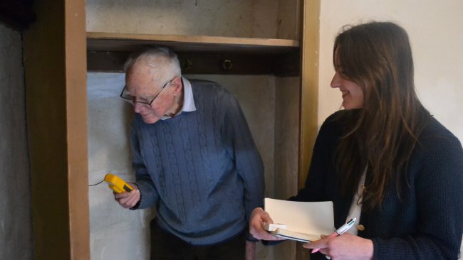 An older man checks a gadget while a woman takes notes.
