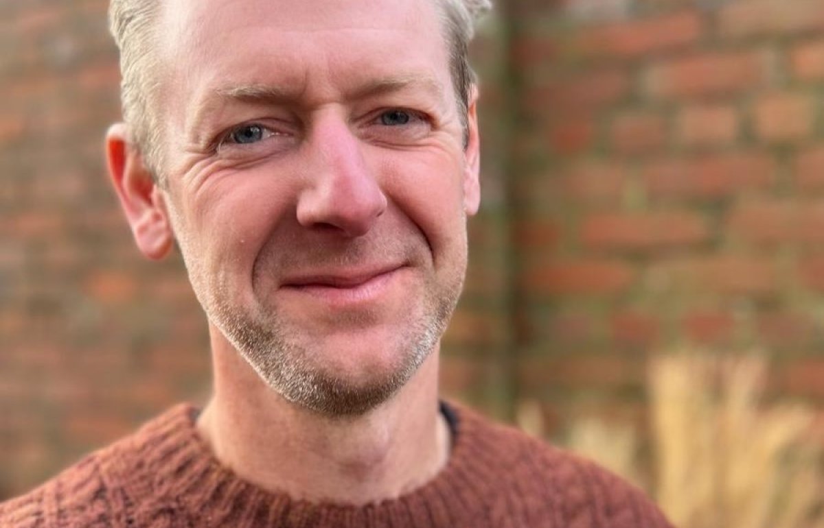 Matt James looking at the camera. Man with grey hair and a brick wall behind