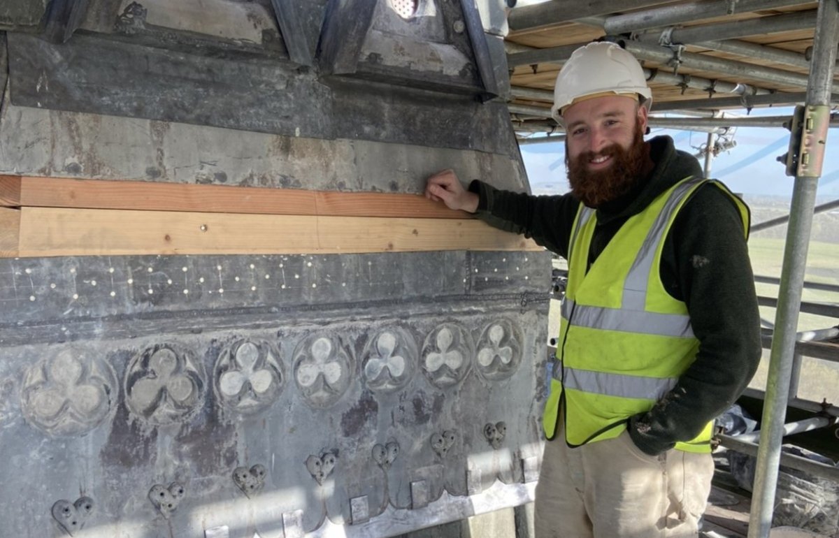 A smiling man with a beard stands on scaffolding in front of historic details. He has a hard hat and high-vis vest.