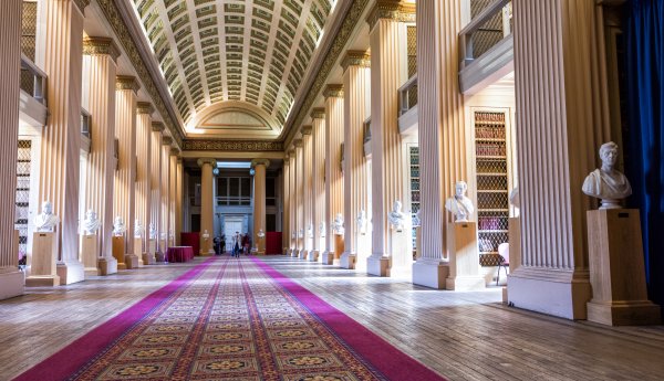 A grand, classical interior hall with tall fluted columns, an ornate arched ceiling, and a long red carpet, lined with marble busts on pedestals leading toward a distant doorway