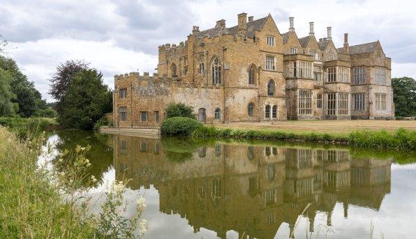 Historic stone manor house beside a calm moat, reflected in the water under a cloudy sky, with trees and grasses surrounding the grounds